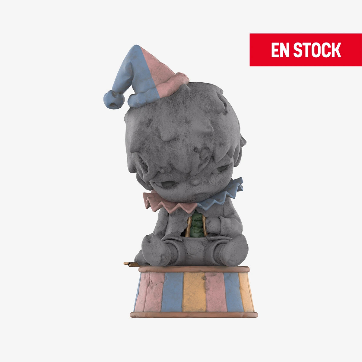 Figura coleccionable de Hirono de la serie Reshape, con apariencia de estatua de piedra deteriorada. Está sentado sobre una base a rayas en tonos pastel azul, rosa y amarillo, con un sombrero de payaso inclinado y cuello de volantes en los mismos colores. La expresión del personaje es melancólica. En la esquina superior derecha aparece una etiqueta roja con el texto 