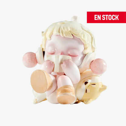 Figura coleccionable de SKULLPANDA de la serie "The Warmth", mostrando a un personaje con los ojos cerrados leyendo un libro, sentado con expresión tranquila y relajada. Viste auriculares con esferas rosadas y está acompañado por un pequeño perrito dormido a su lado. En la esquina superior derecha aparece una etiqueta roja con el texto "EN STOCK"